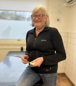 Kvinna med glasögon, ljust hår och mörk tröja halvsitter på ett undersökningsbord. Hon håller trimsaxar i händerna.